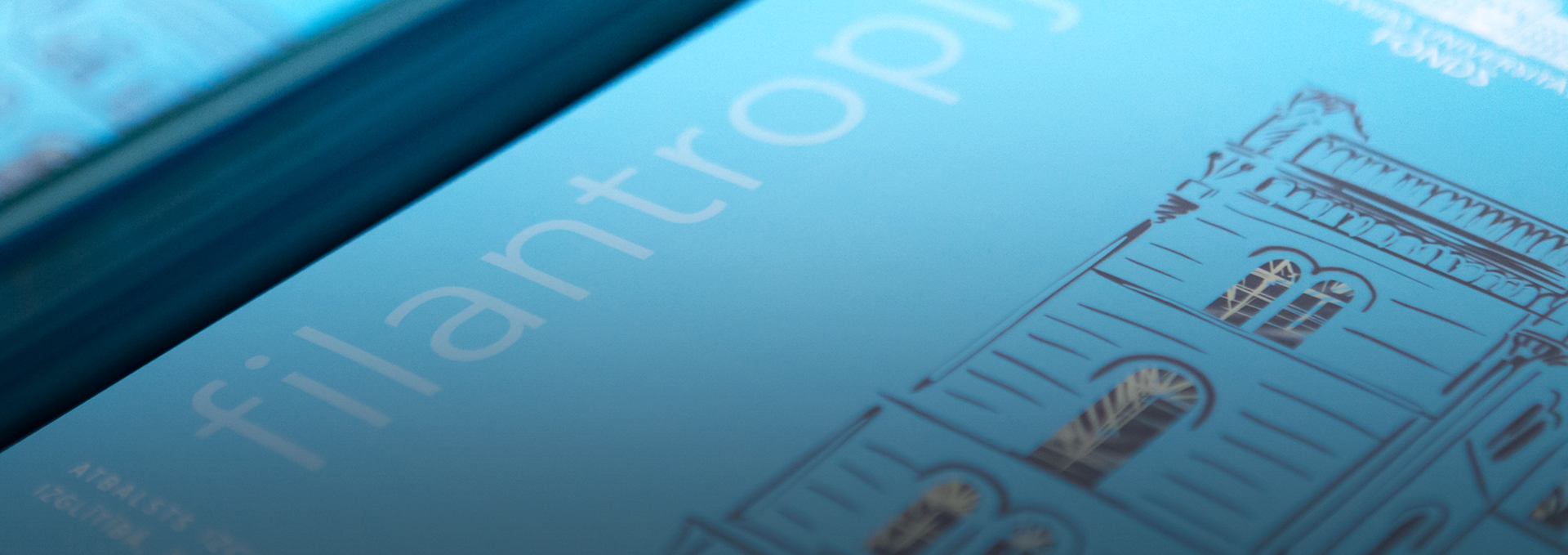 A close-up, angled view of a the word 'filantropi' and the blurred image of an architectural drawing, with a blue tone overlay.
