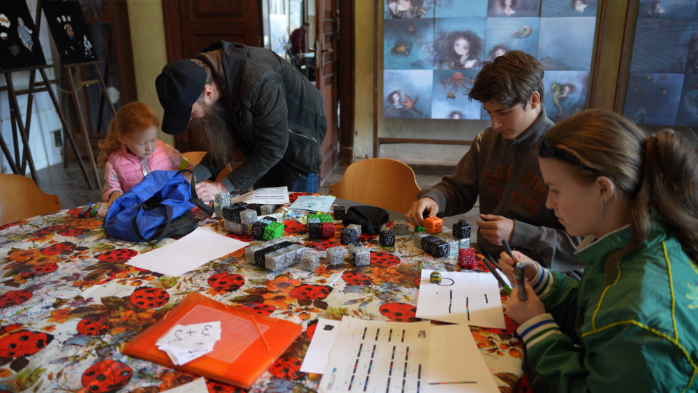 A group of children engaged in creative activities at a table covered with a ladybug-patterned cloth. An adult, bending over, appears to assist a young girl with her work, while two other children are focused on their own crafting, surrounded by colorful blocks and art materials.
