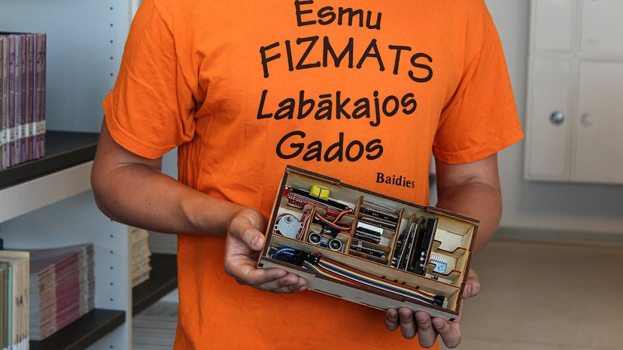 A person in an orange shirt with text 'Esmu FIZMATS Labākajos Gados' holds a wooden box filled with neatly arranged electronic components and tools.